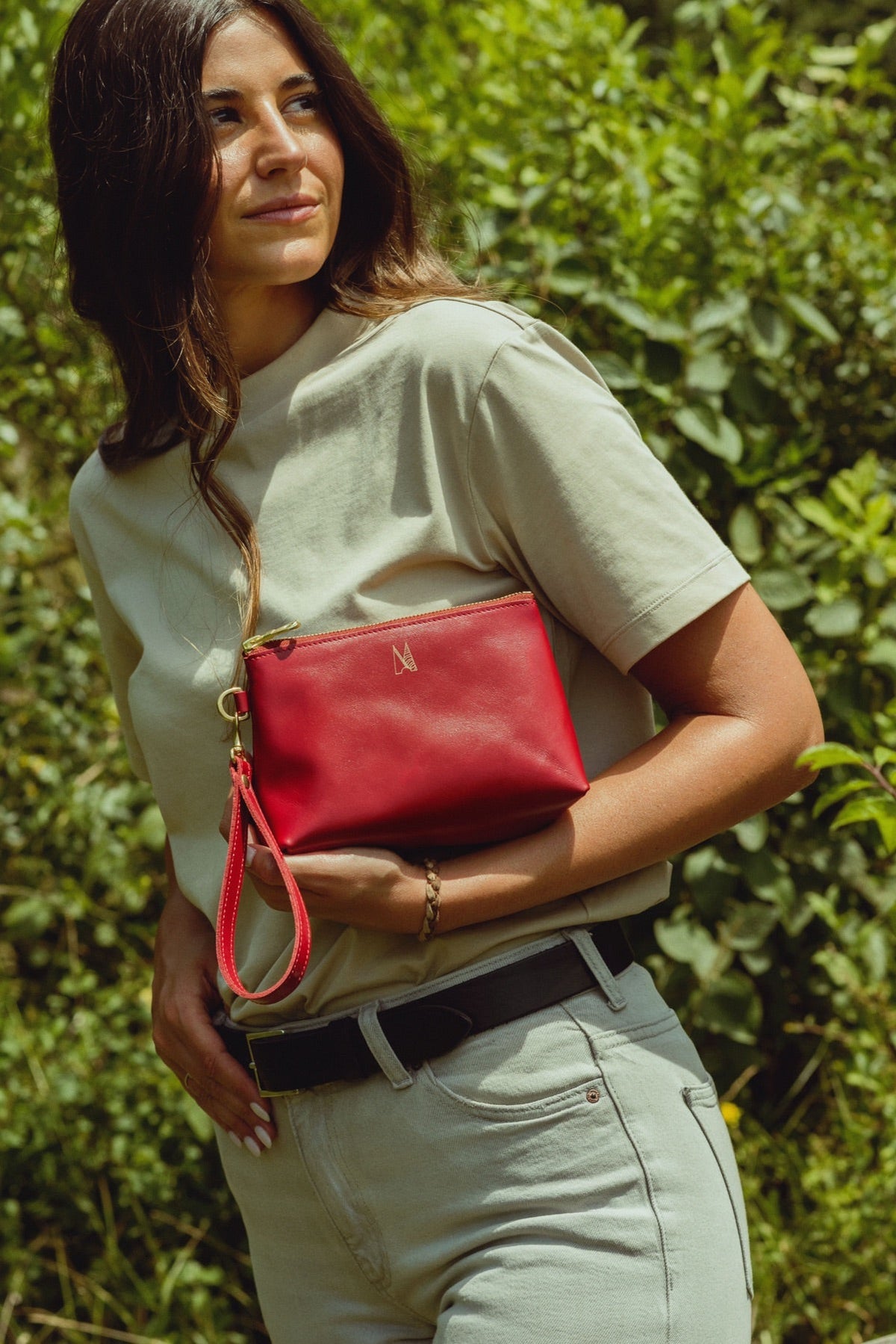 Woman holding a red leather zip pouch with wrist strap against her waist, glancing to one side